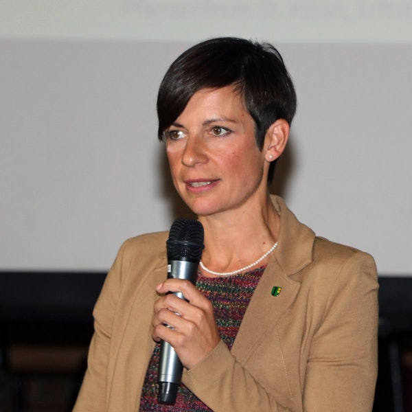 Zu sehen ist eine Frau mit dunklen Haaren vor einem hellen Hintergrund.