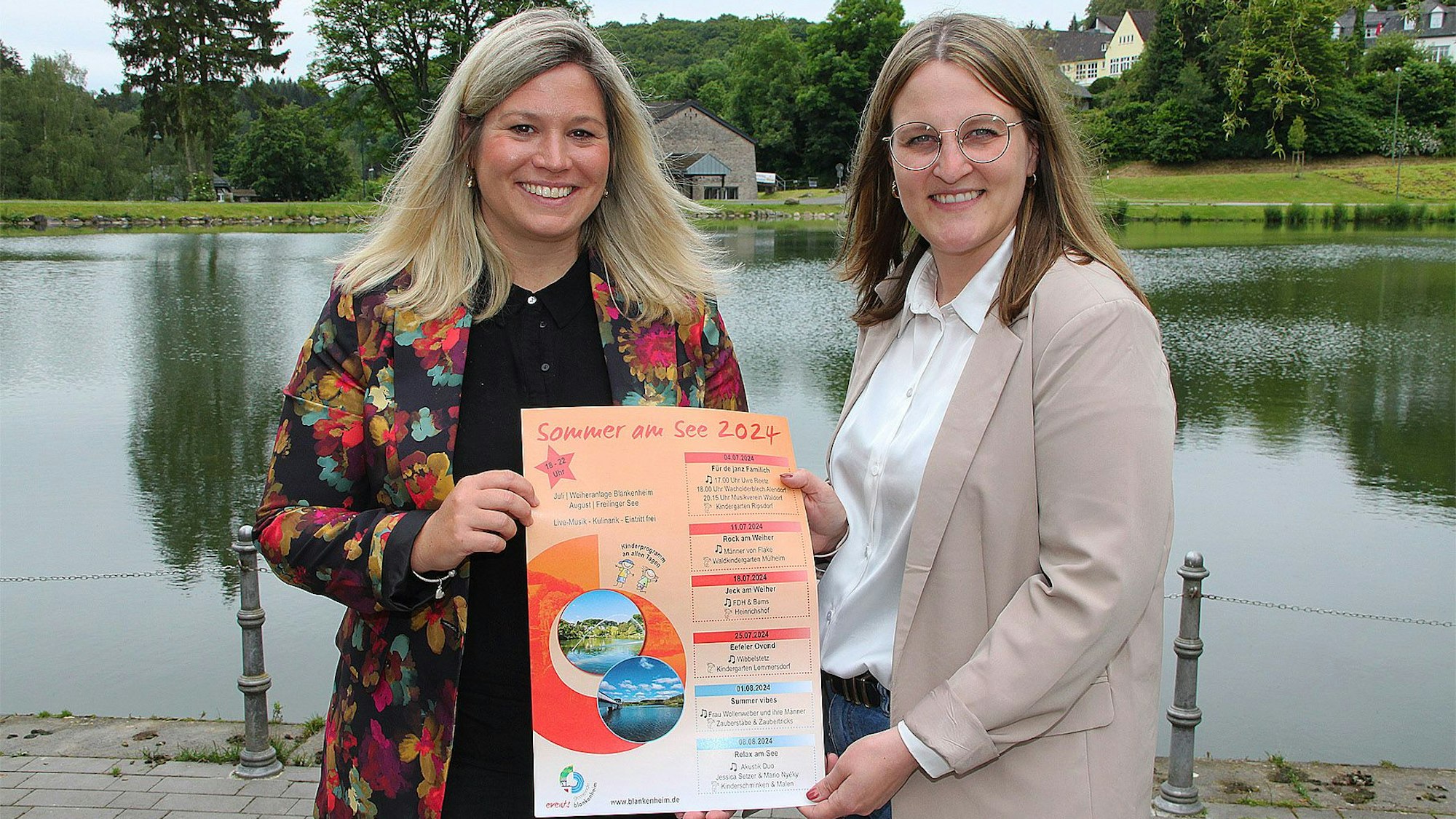 Bürgermeisterin Jennifer Meuren (l.) und Touristikerin Christina Gier stehen an der Weiheranlage in Blankenheim. Sie halten ein Plakat der Konzertreihe „Sommer am See“ in den Händen.