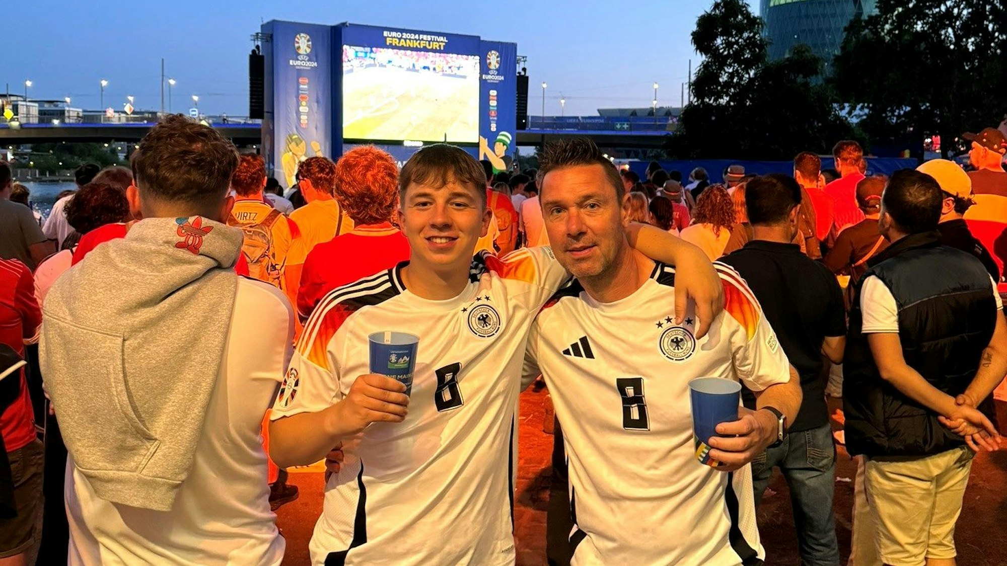 Uwe und Luis Metternich tragen beide das Toni-Kroos-Trikot und halten einen Bierbecher in der Hand. Im Hintergrund die Leinwand beim Public Viewing in Frankfurt.