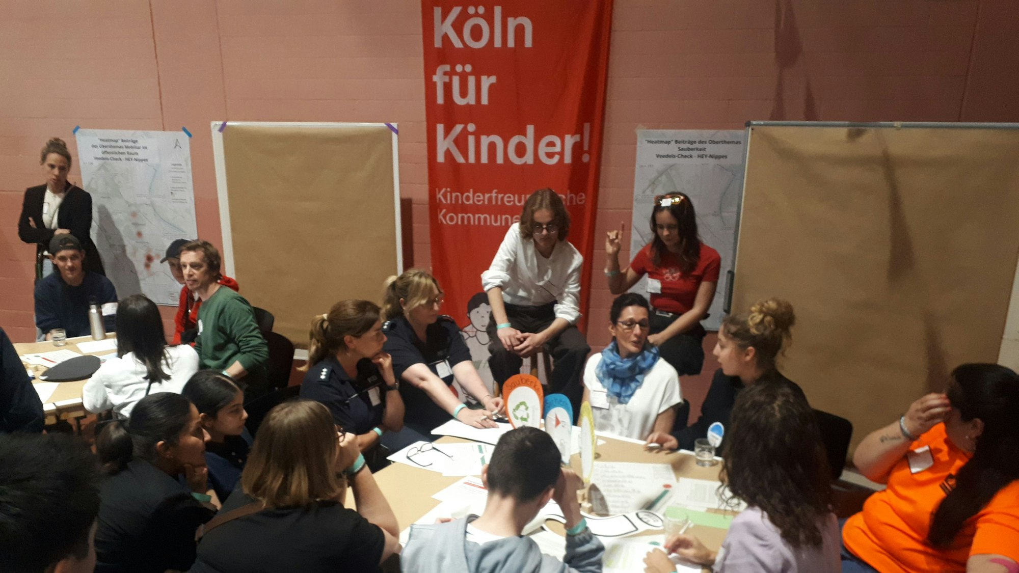 Zu sehen sind Kinder, Jugendliche und Erwachsene, die an mehreren Tischen diskutieren.
