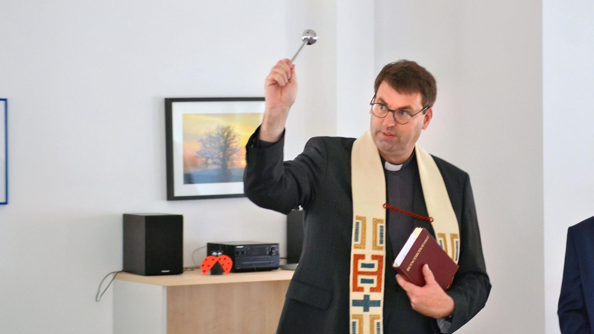 Kreisdechant Guido Zimmermann trägt einen schwarzen Anzug und helle Stola. Er hält ein Aspergill in der Hand und segnet ein Zimmer der Caritas-Tagespflege in Euskirchen ein.