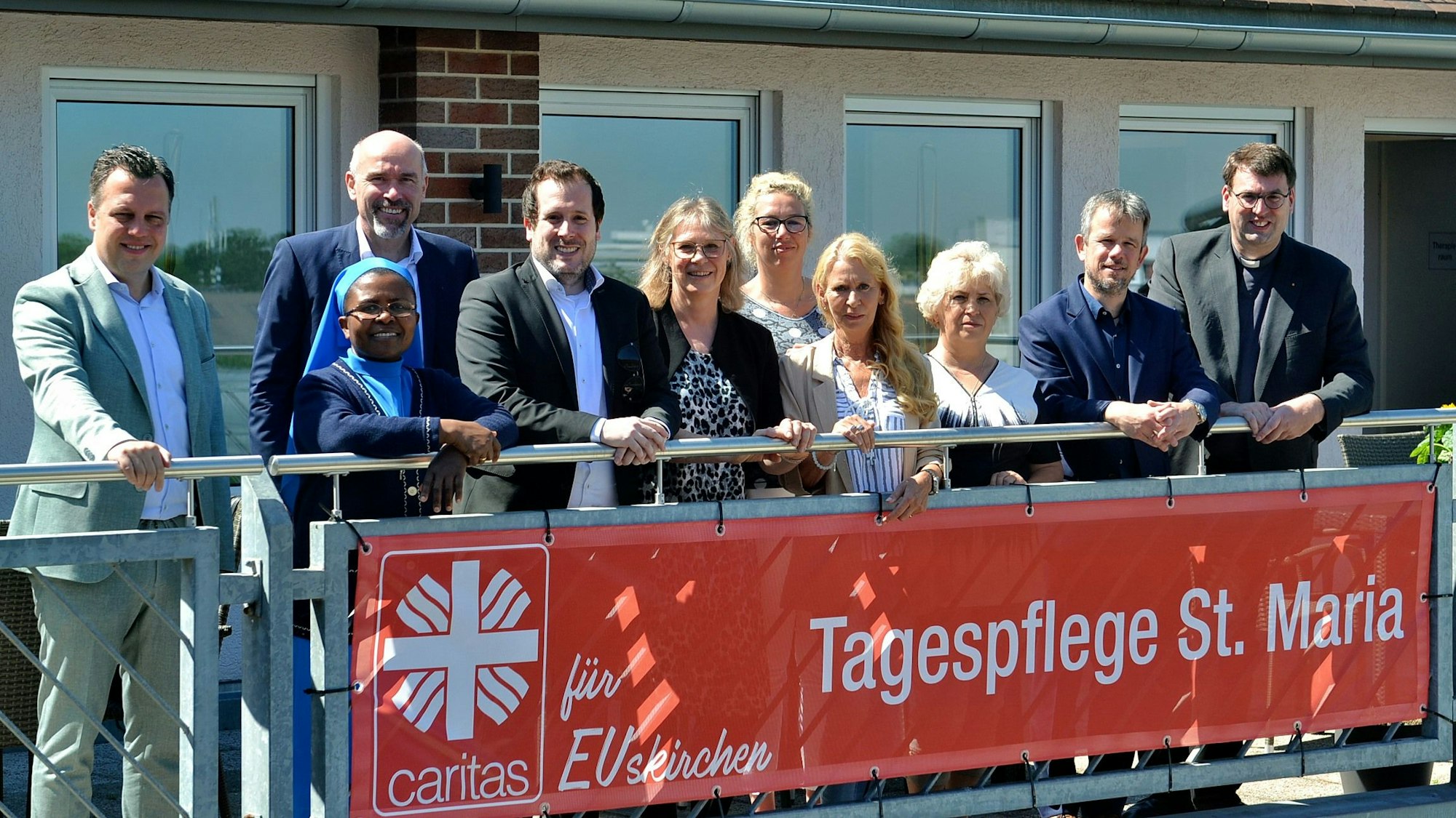 Einige Frauen und Männer stehen auf einem Balkon in Euskirchen, an dem ein rotes Banner hängt, auf dem das Caritas-Symbol zu sehen ist und auf dem steht „Tagespflege St. Maria“.