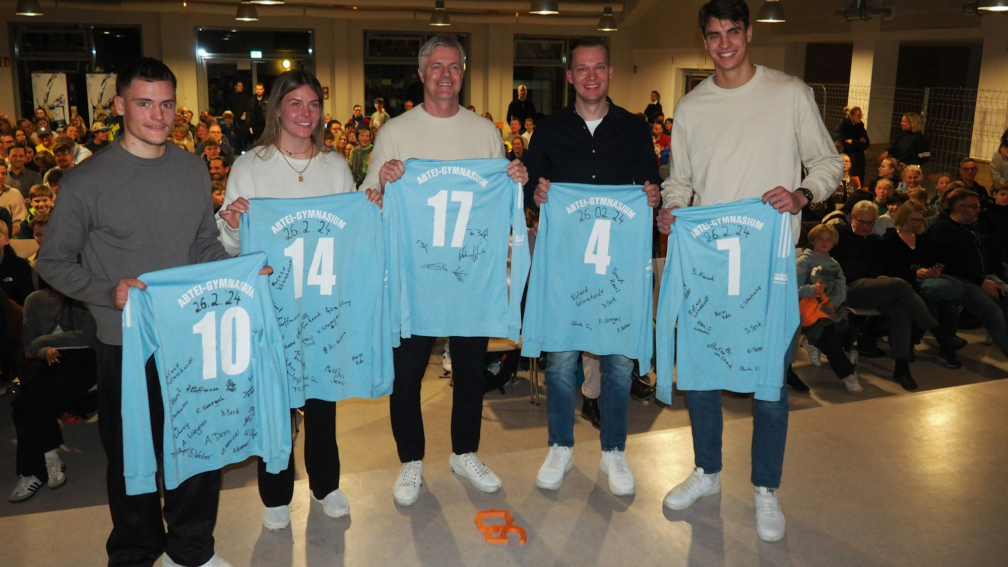 Eine Frau und vier Männer lächeln in die Kamera und zeigen T-Shirts, die ihre ehemaligen Lehrerinnen und Lehrer unterschrieben haben.