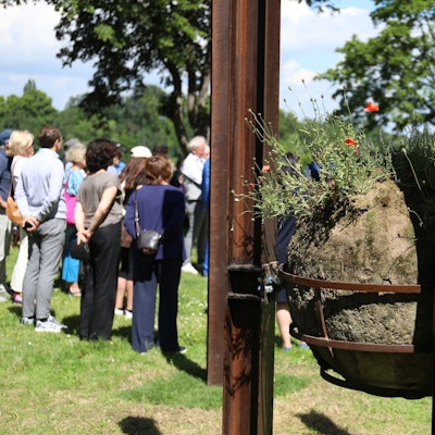 In einem Metallkorb, der an Stahlstelen befestigt ist, liegt ein großer Lehmball. Aus ihm sprießen Mohnblumen.