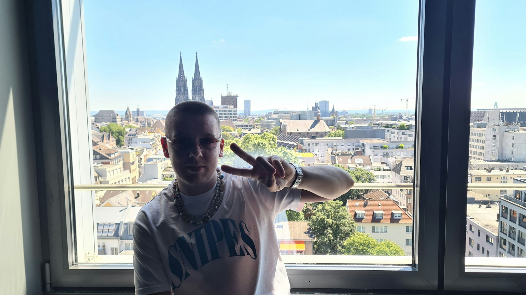 Der britische Rapper Aitch posiert vor einem Fenster, dadurch ist im Hintergrund der Kölner Dom zu sehen.