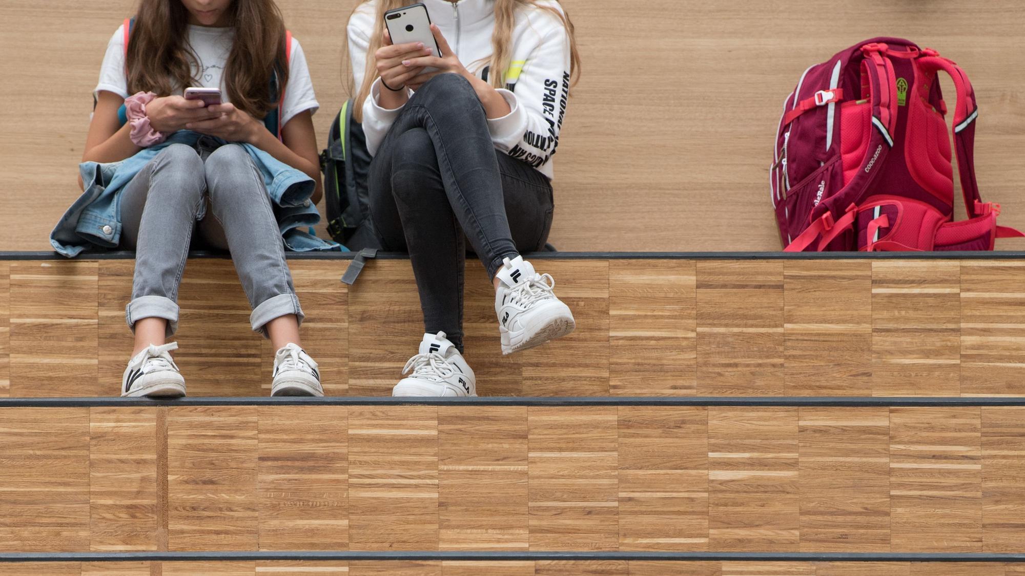 Schülerinnen sitzen im Foyer eines Gymnasium auf einer Freitreppe und halten jeweils ein Smartphone. Das Handyverbot an bayerischen Schulen soll gelockert werden: Künftig sollen die Schulen selber entscheiden dürfen, ob sie Schülerinnen und Schülern die private Handynutzung in den Pausen oder über Mittag erlauben oder nicht. (zu dpa "Handyverbot an Schulen wird gelockert - Entscheidung künftig vor Ort") +++ dpa-Bildfunk +++