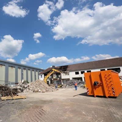 Neben einem Fabrikgebäude stehen ein Bagger und ein Container, am Rand liegt Bauschutt.