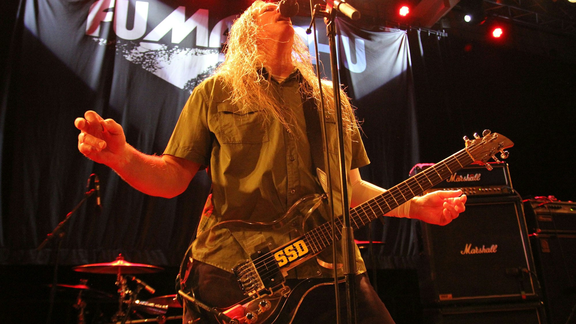 Der Sänger und Gitarrist Scott Hill steht singend am Mikrofon und breitet die Arme aus. Im Hintergrund prangt der Schriftzug der Band auf einem schwarzen Vorhang.