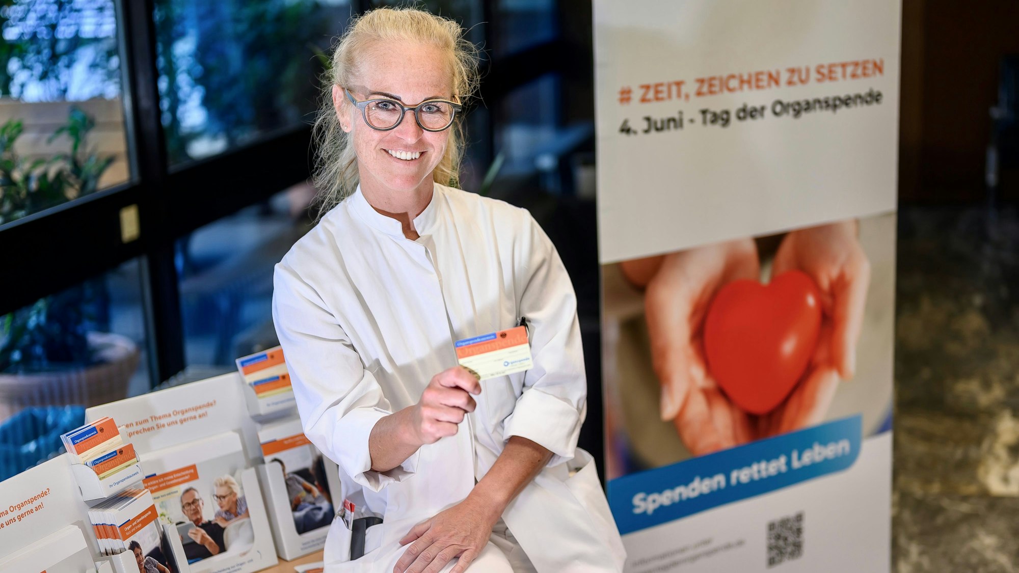 Eine junge Ärztin sitzt auf einem Tisch und hält lächelnd einen Organspendeausweis in die Kamera. Auf dem Tisch befinden sich Ständer mit Informationsmaterial zur Organspende, rechts daneben steht ein Aufsteller, der für den „4. Juni – Tag der Organspende“ wirbt.