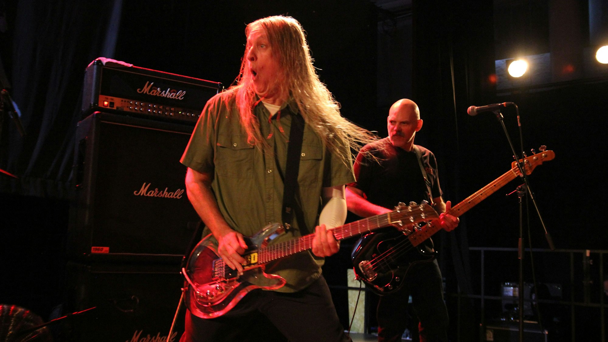 Ein Gitarrist steht vor dem Bassisten. Hinter beiden sind aufgetürmte Verstärker zu sehen.