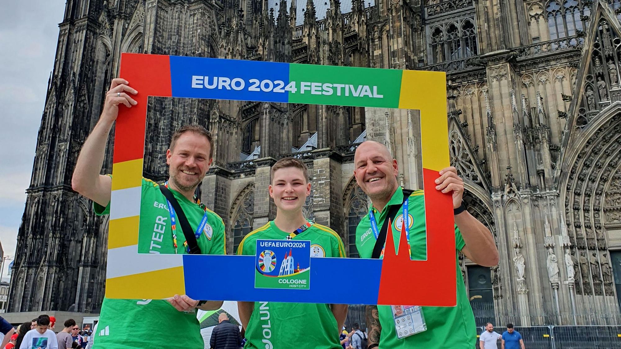 Die EM-Volunteers stehen vor dem Kölner Dom und halten einen Rahmen mit der Aufschrift „Euro 2024 Festival“.