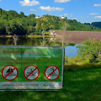 Vor dem Kronenburger See ist ein Schild aufgestellt. Die Aufschrift lautet: „Aufgrund technischer Arbeiten nach dem Hochwasser 2021 ist eine Nutzung der Wasserfläche in diesem Jahr leider nicht gestattet!“