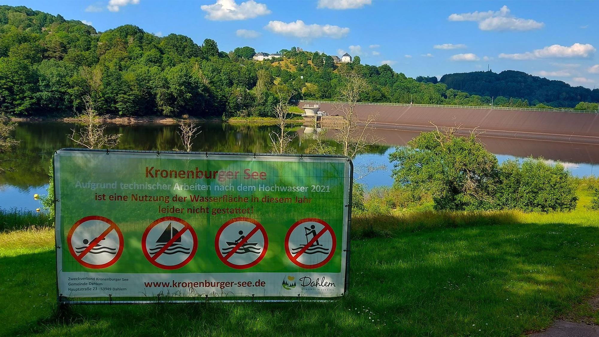 Vor dem Kronenburger See ist ein Schild aufgestellt. Die Aufschrift lautet: „Aufgrund technischer Arbeiten nach dem Hochwasser 2021 ist eine Nutzung der Wasserfläche in diesem Jahr leider nicht gestattet!“