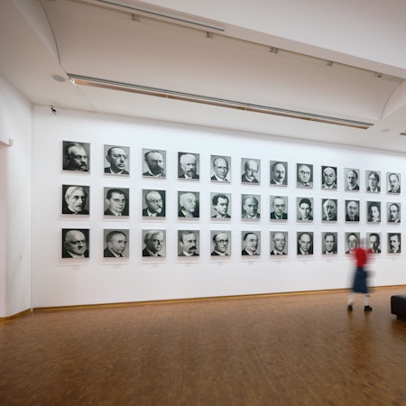 Eine Frau geht vor einer Wand mit 48 Bildern von Männern vorbei.