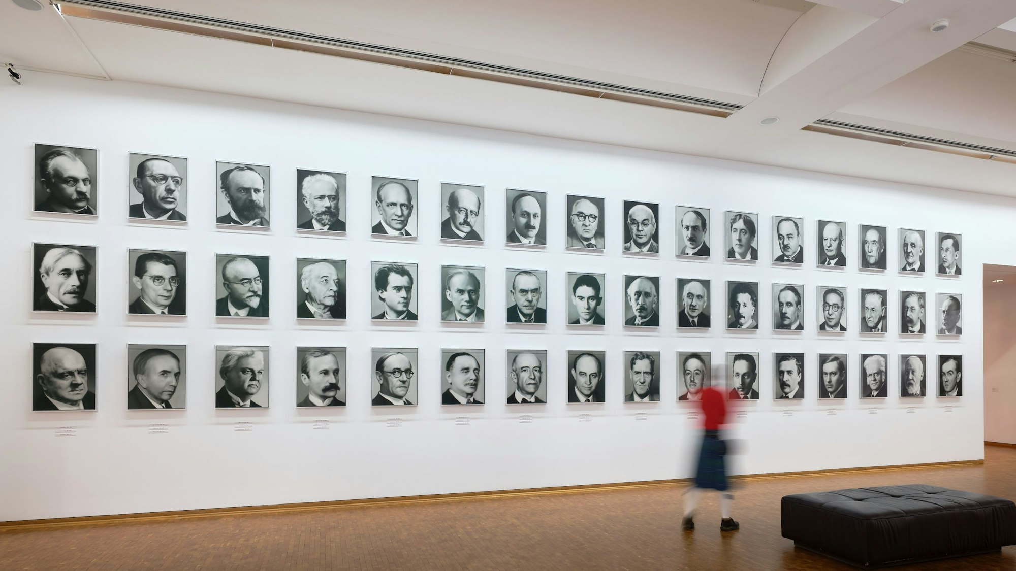 Eine Frau geht vor einer Wand mit 48 Bildern von Männern vorbei.