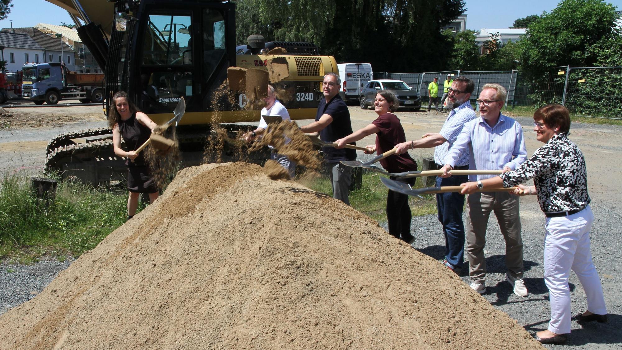 Drei Frauen und vier Männer schaufeln Sand aus einem Sandhaufen, im Hintergrund ein Bagger
