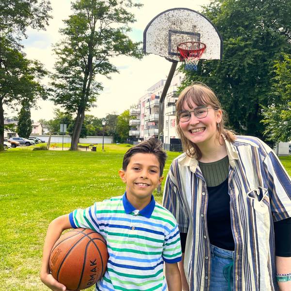 Jusef, 10, aus Sysien steht mit seiner Patin Kaja auf einem Spielplatz in Köln Höhenhaus. Er trägt einen Basketball unter dem Arm, im Hintergrund ist ein Basketballkorb zu sehen.