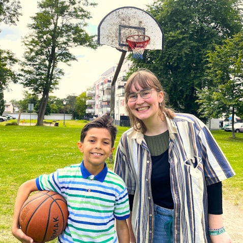 Jusef, 10, aus Sysien steht mit seiner Patin Kaja auf einem Spielplatz in Köln Höhenhaus. Er trägt einen Basketball unter dem Arm, im Hintergrund ist ein Basketballkorb zu sehen.