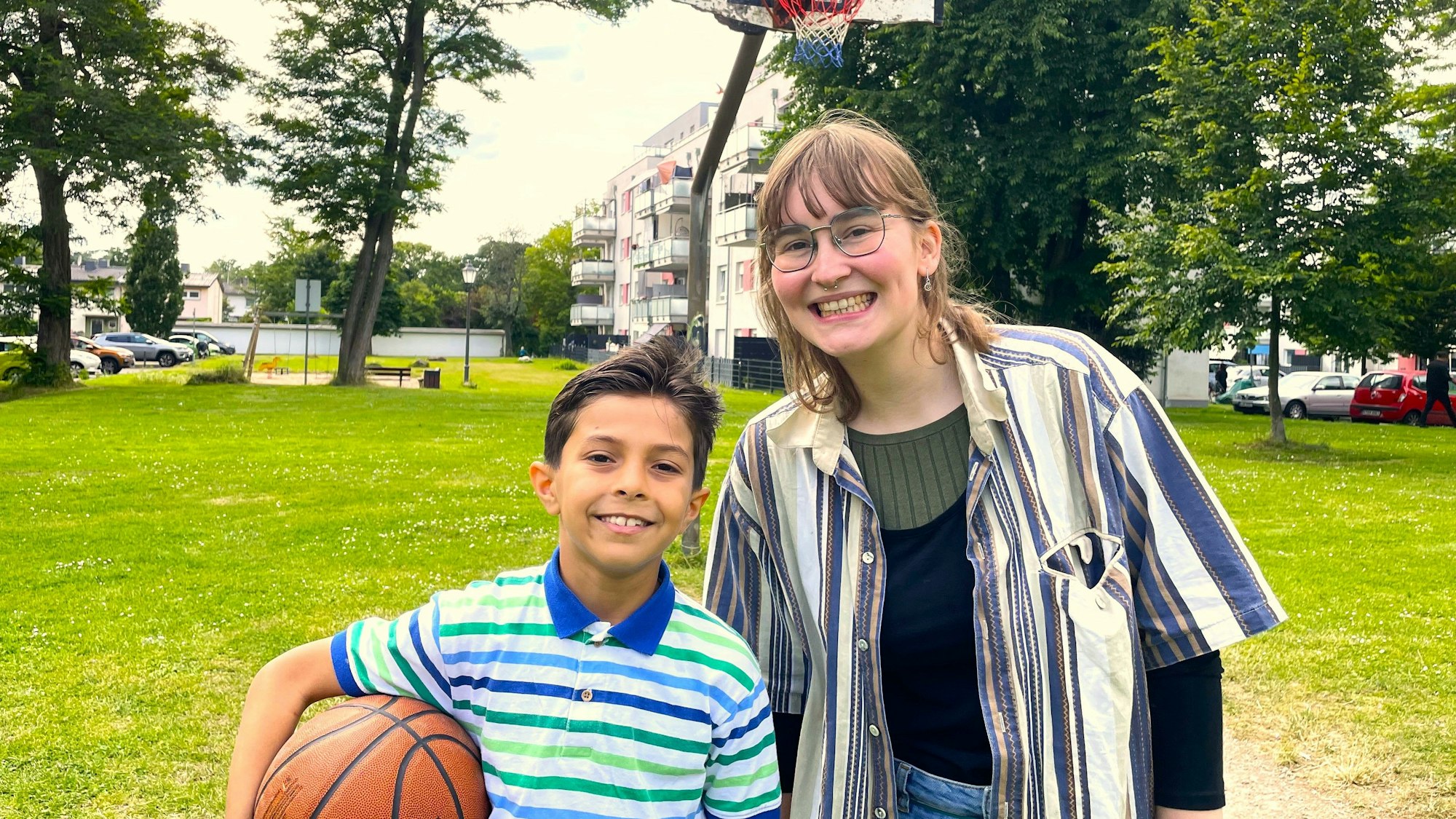 Jusef, 10, aus Sysien steht mit seiner Patin Kaja auf einem Spielplatz in Köln Höhenhaus. Er trägt einen Basketball unter dem Arm, im Hintergrund ist ein Basketballkorb zu sehen.