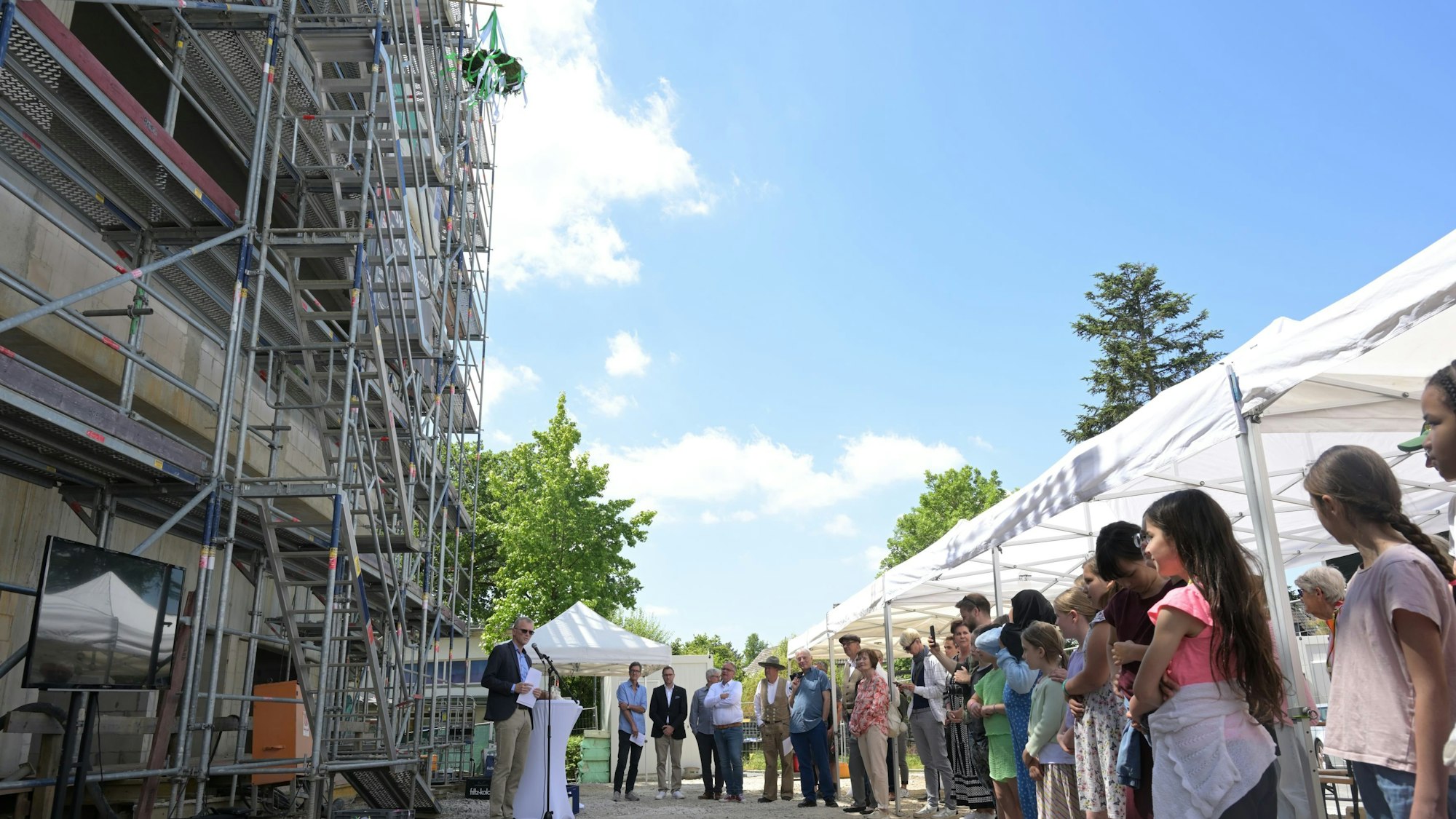 Links ist ein Teil des eingerüsteten Rohbaus zu sehen. Rechts ein Teil der Gäste. Der Bürgermeister spricht in ein Mikrophon.