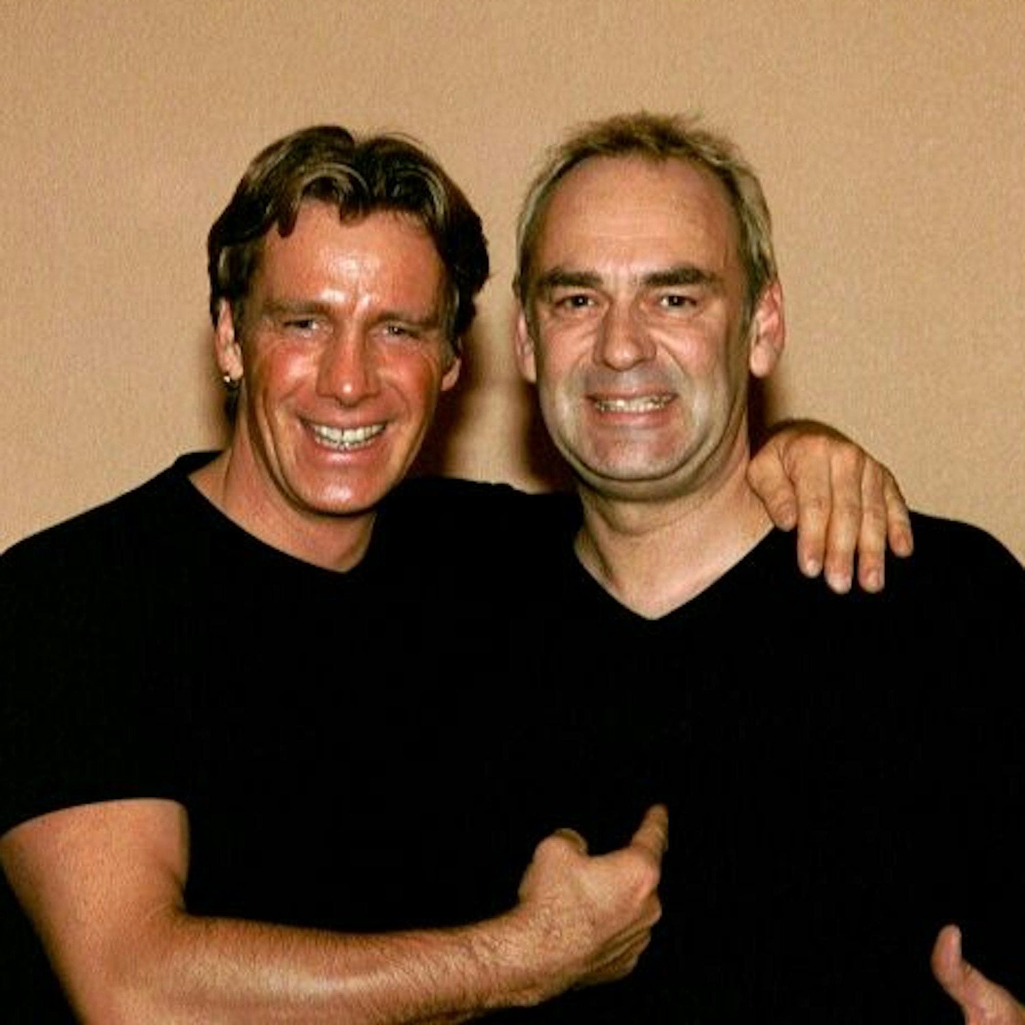 Zwei Männer in schwarzen T-Shirts stehen Arm in Arm nebeneinander.