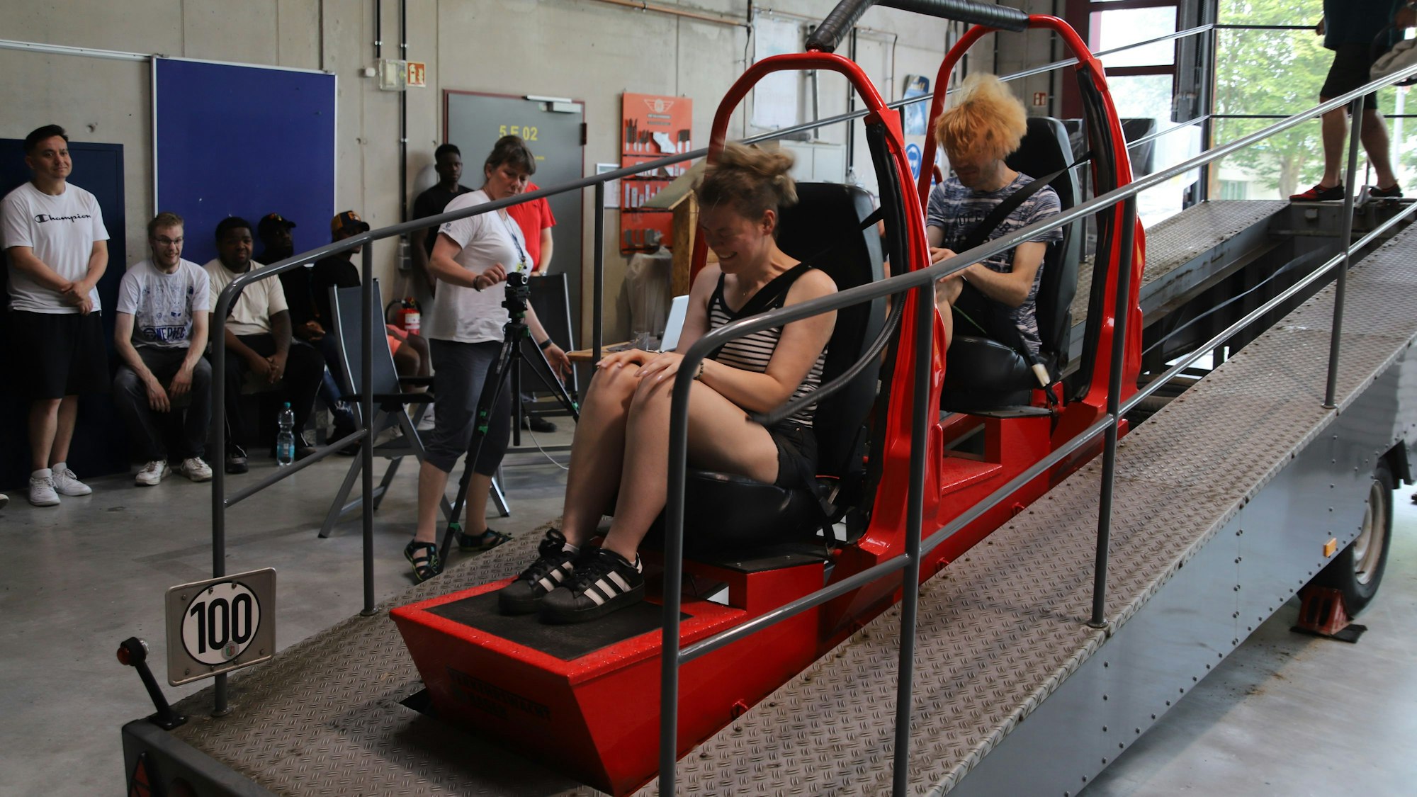 Ein Gurtschlitten fährt nach unten, zwei Menschen werden im Augenblick des Aufpralls nach vorne geschoben.