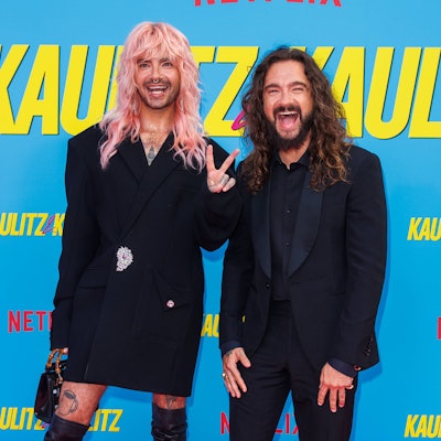 Bill und Tom Kaulitz stehen vor einer Wand mit dem Slogan ihrer Netflix-Doku und grüßen in die Kamera
