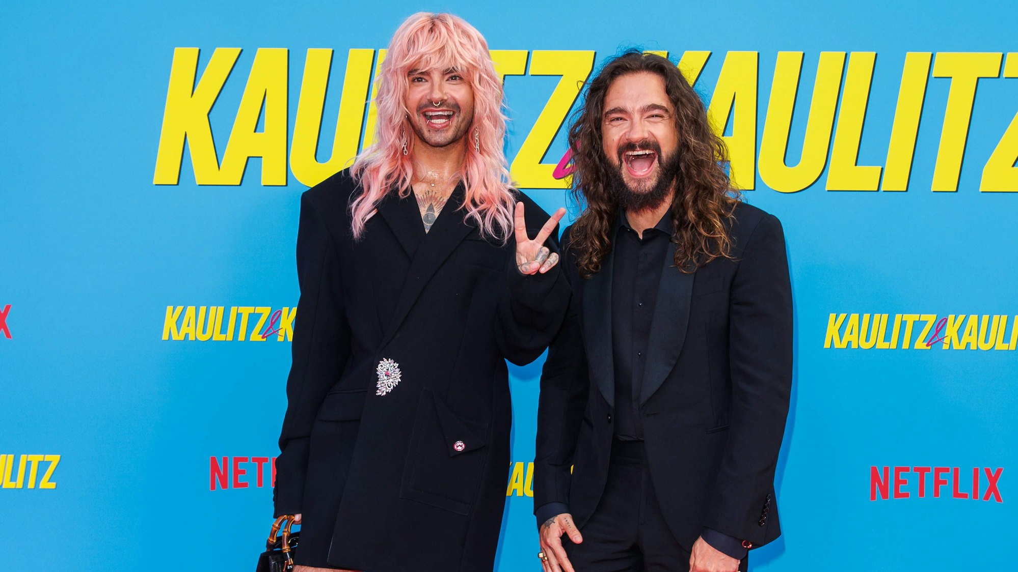 Bill und Tom Kaulitz stehen vor einer Wand mit dem Slogan ihrer Netflix-Doku und grüßen in die Kamera