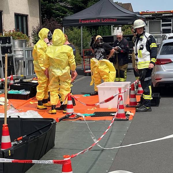 Mehrere Einsatzkräfte in gelben Schutzanzügen stehen auf einer abgesperrten Fläche vor einem Mehrfamilienhaus in Lohmar.