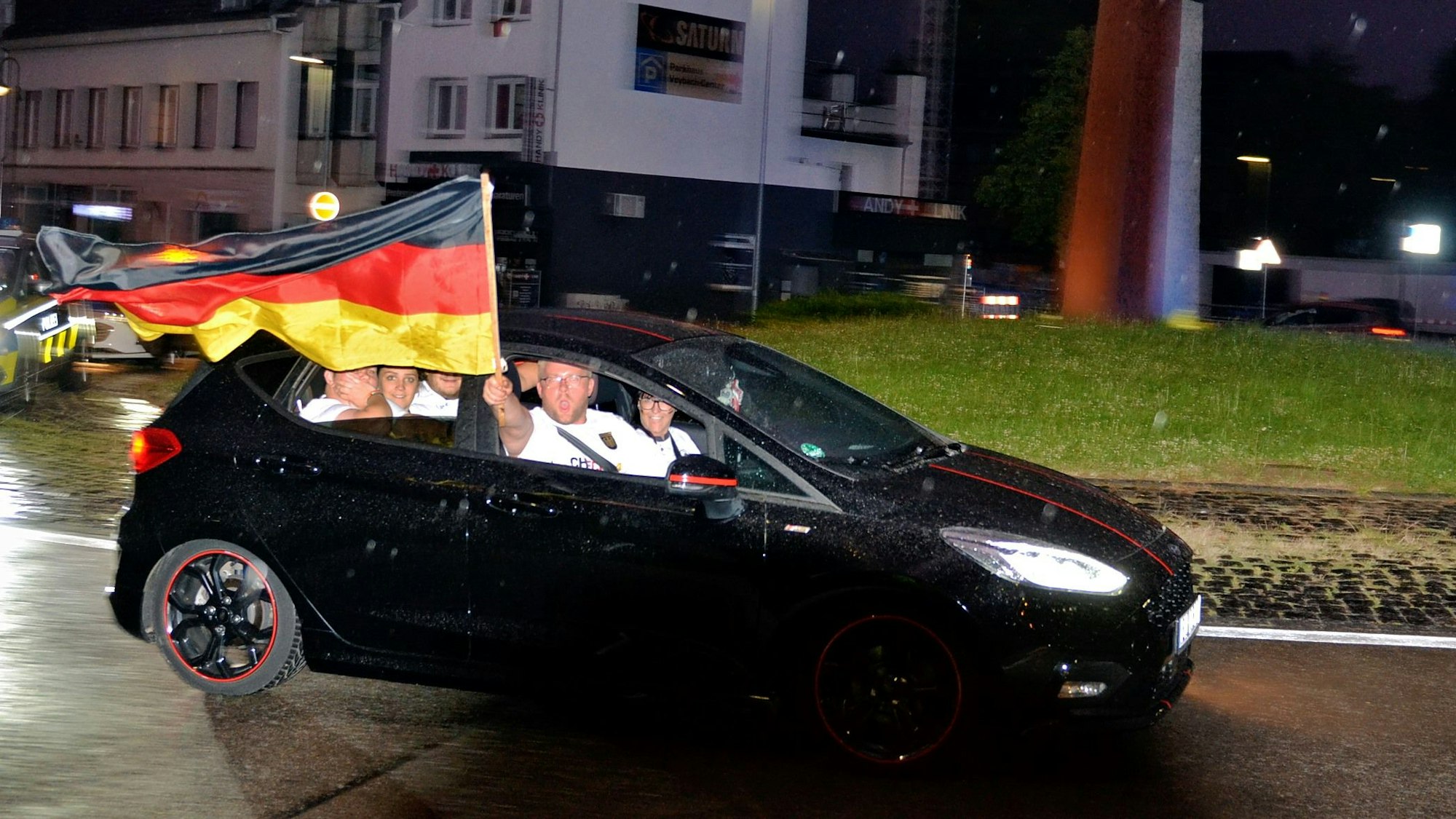 Fünf Fußballfans sitzen in einem schwarzen Ford Fiesta. Einer hält eine Deutschland-Fahne aus dem offenen Fenster des fahrenden Autos.
