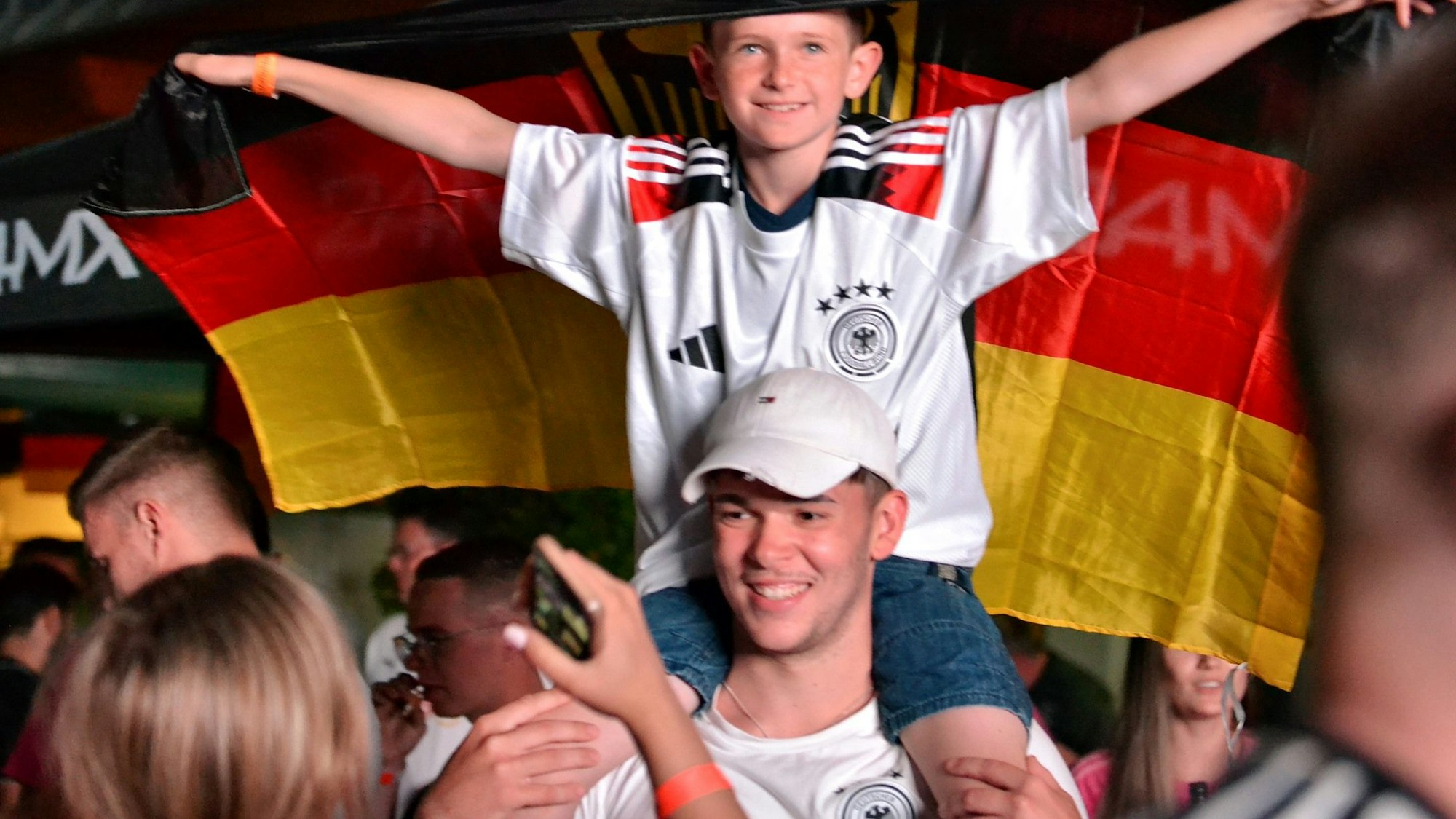 Ein kleiner Junge im Deutschland-Trikot hält eine Deutschland-Fahne hinter sich in die Höhe und sitzt auf den Schultern eines jungen Mannes.