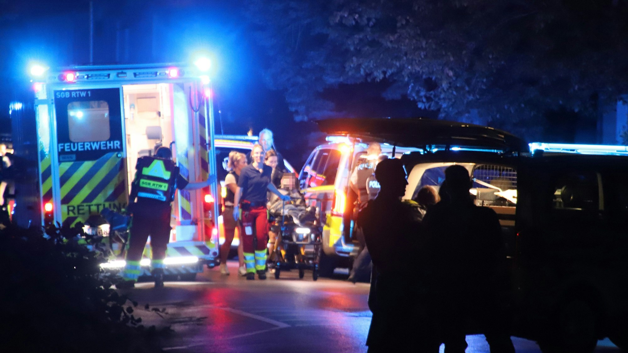 Mehrere Einsatzfahrzeuge von Polizei und Feuerwehr stehen auf einer nächtlichen Straße, Rettungskräfte schieben eine Trage.