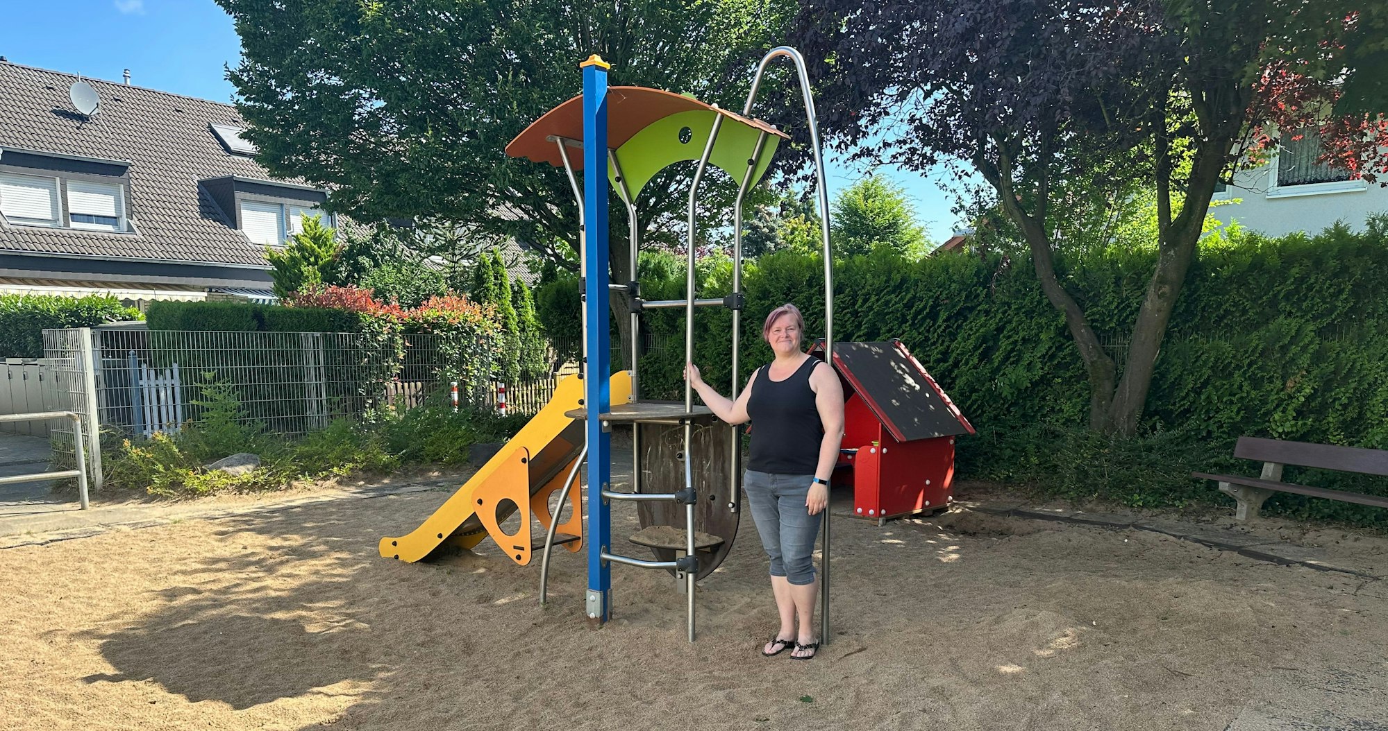 Eine Frau in sommerlicher Kleidung steht an einem Klettergerüst mit gelber Rutsche. Im Hintergrund ist eine kleine rote Hütte zu sehen.