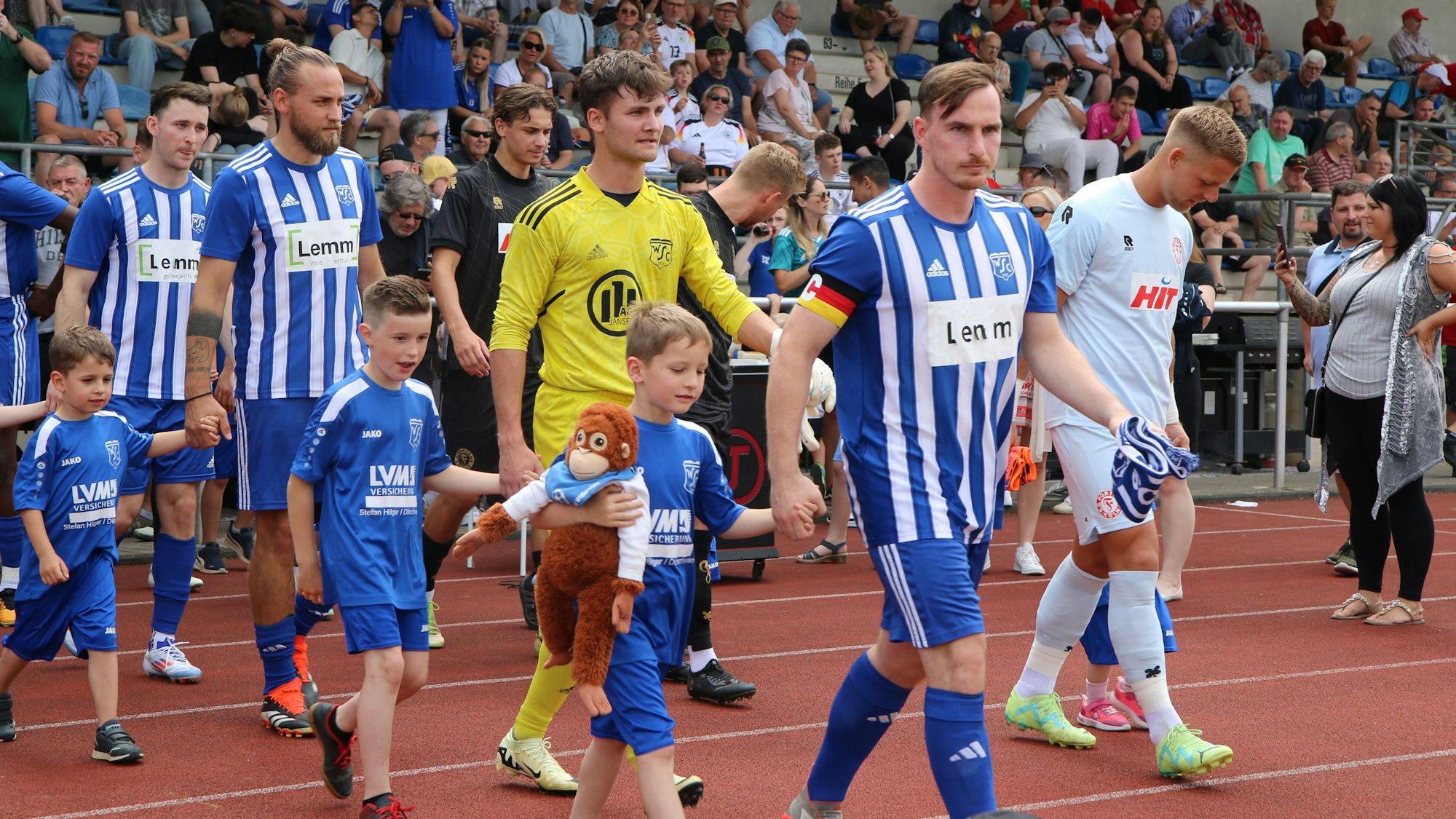 Die Mannschaften gehen aufs Spielfeld. An den Händen halten die Spieler Einlaufkinder. Eines trägt ein Stofftier.