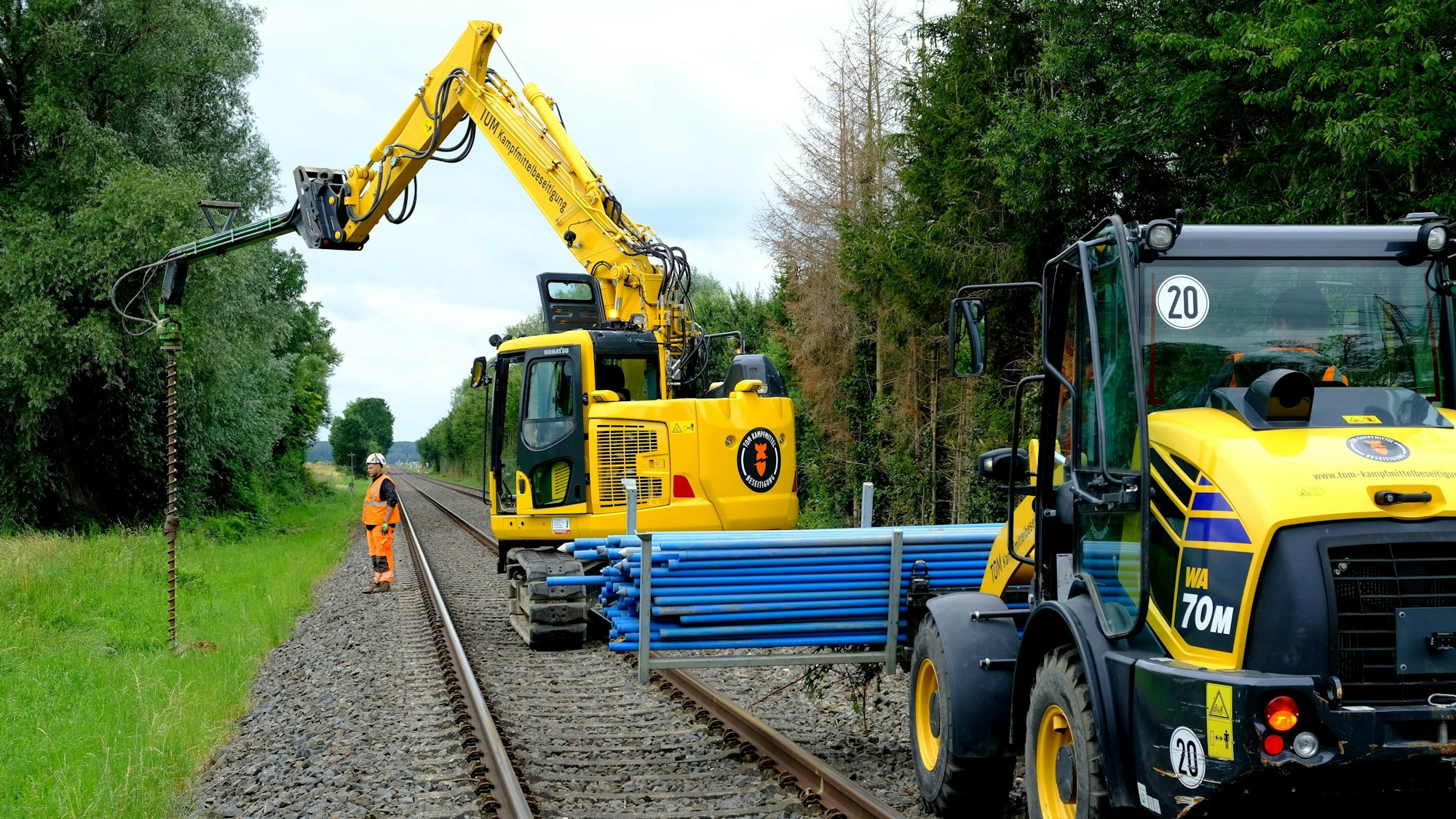 Mitarbeiter eines Kampfmittelräumers sind im Bereich Euskirchen/Euenheim mit Vorsondierungen an den künftigen Standorten für die Oberleitungsmasten beschäftigt. An einem Bagger ist ein Bohrgestänge befestigt, auf der Schaufel eines Radladers wird Material transportiert.