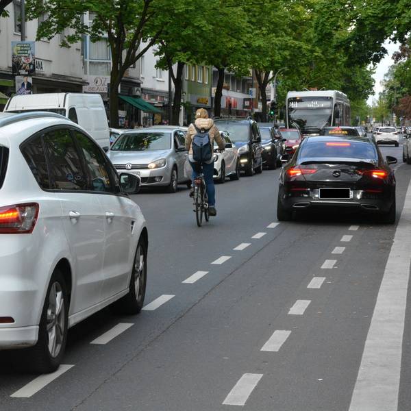 Ein Auto parkt auf einem Radweg, eine Radfahrerin überholt in zweiter Reihe. Ihr folgt dicht ein Auto.