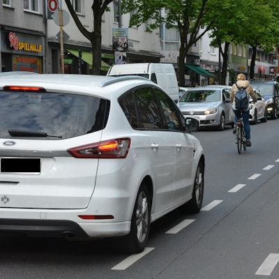 Ein Auto parkt auf einem Radweg, eine Radfahrerin überholt in zweiter Reihe. Ihr folgt dicht ein Auto.