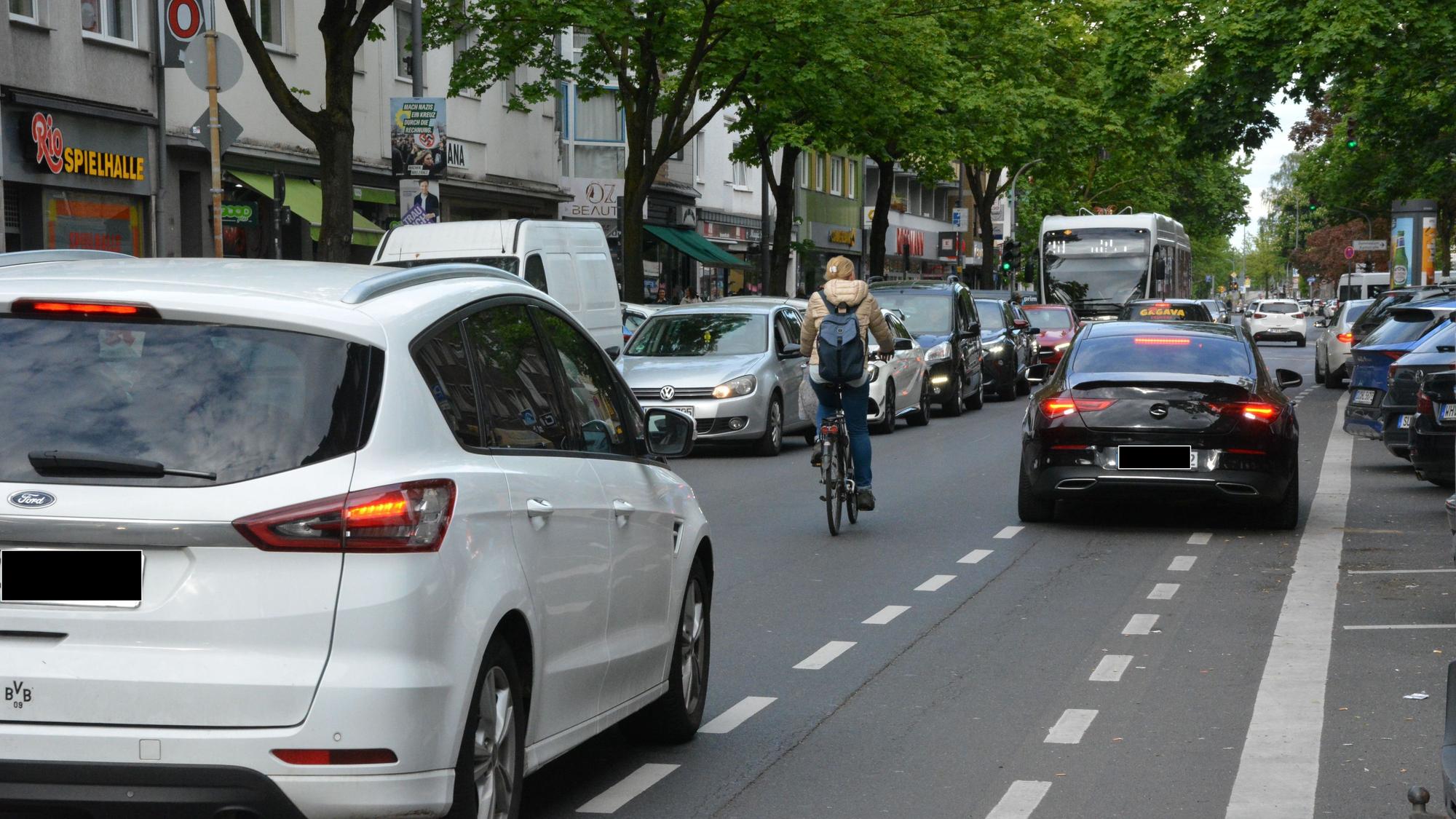 Ein Auto parkt auf einem Radweg, eine Radfahrerin überholt in zweiter Reihe. Ihr folgt dicht ein Auto.