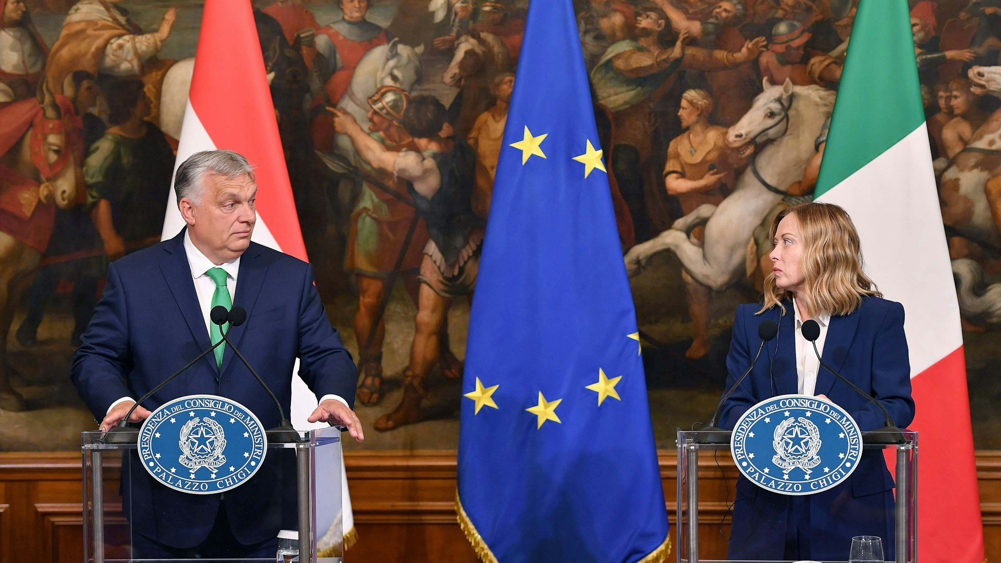 Orban und Meloni stehen an Pulten, sie haben Flaggen ihrer Länder im Hintergrund. Zwischen ihnen hängt die europäische Fahne.