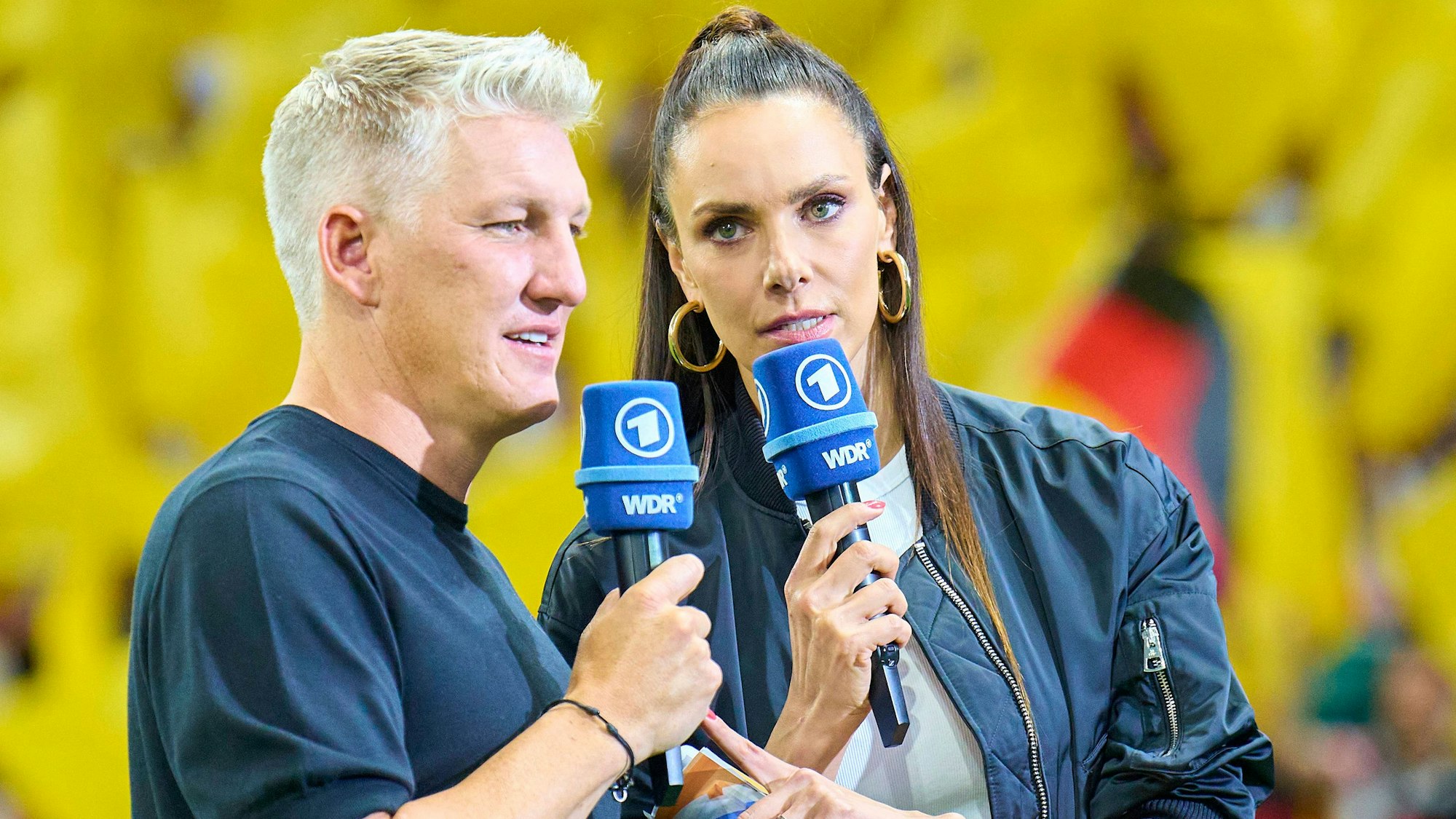 Die TV-Sportmoderatorin Esther Sedlaczek steht neben Bastian Schweinsteiger, Ex-Fußballer und TV-Experte. Beide halten Mikrofone des WDR in ihrer rechten Hand. Die Beiden stehen vor einem gelben Hintergrund, vor dem unscharf eine Deutschland-Fahne zu erkennen ist.