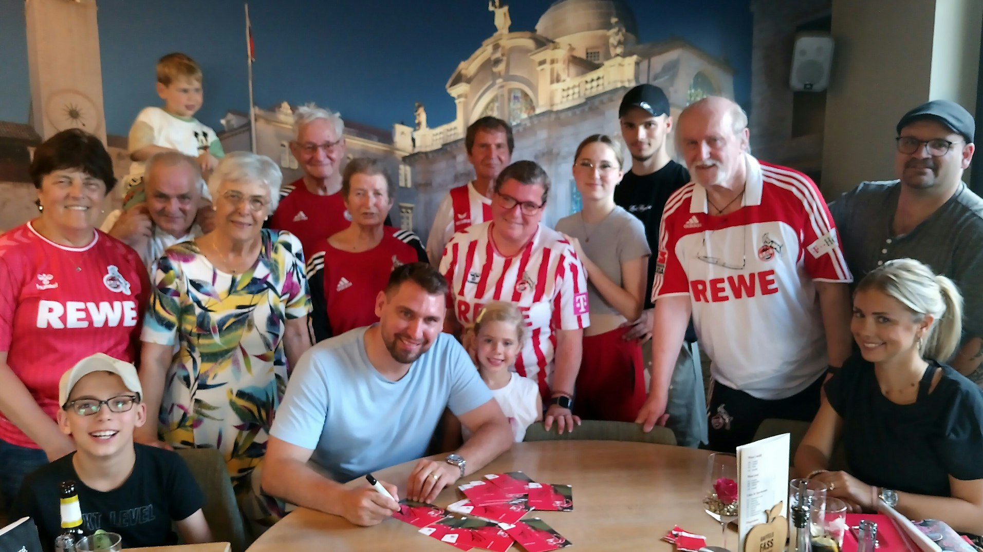 Patrick Helmes sitzt schreibend an einem Tisch, umringt von Mitgliedern des Fan-Clubs. Alle lachen in die Kamera.