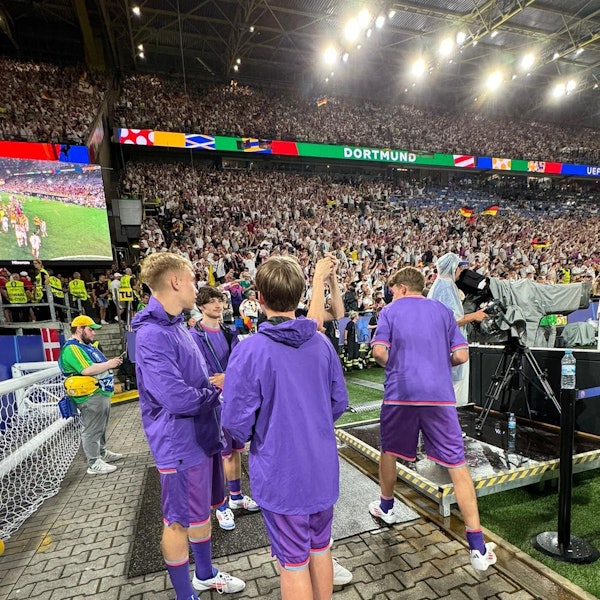 Julian Seliger aus Kirspenich war beim EM-Spiel Deutschland gegen Dänemark als Balljunge dabei. Auf dem Bild ist er links mit drei anderen Balljungen in lila Shirts vor den vollen Rängen des Stadions in Dortmund zu sehen.
