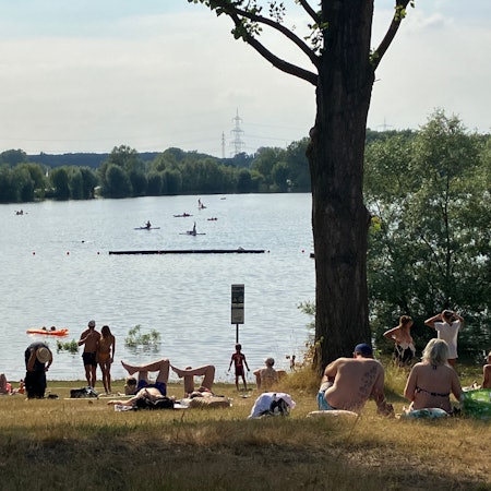 Badegäste sonnen sich an einem Seeufer. Andere schwimmen im Wasser.