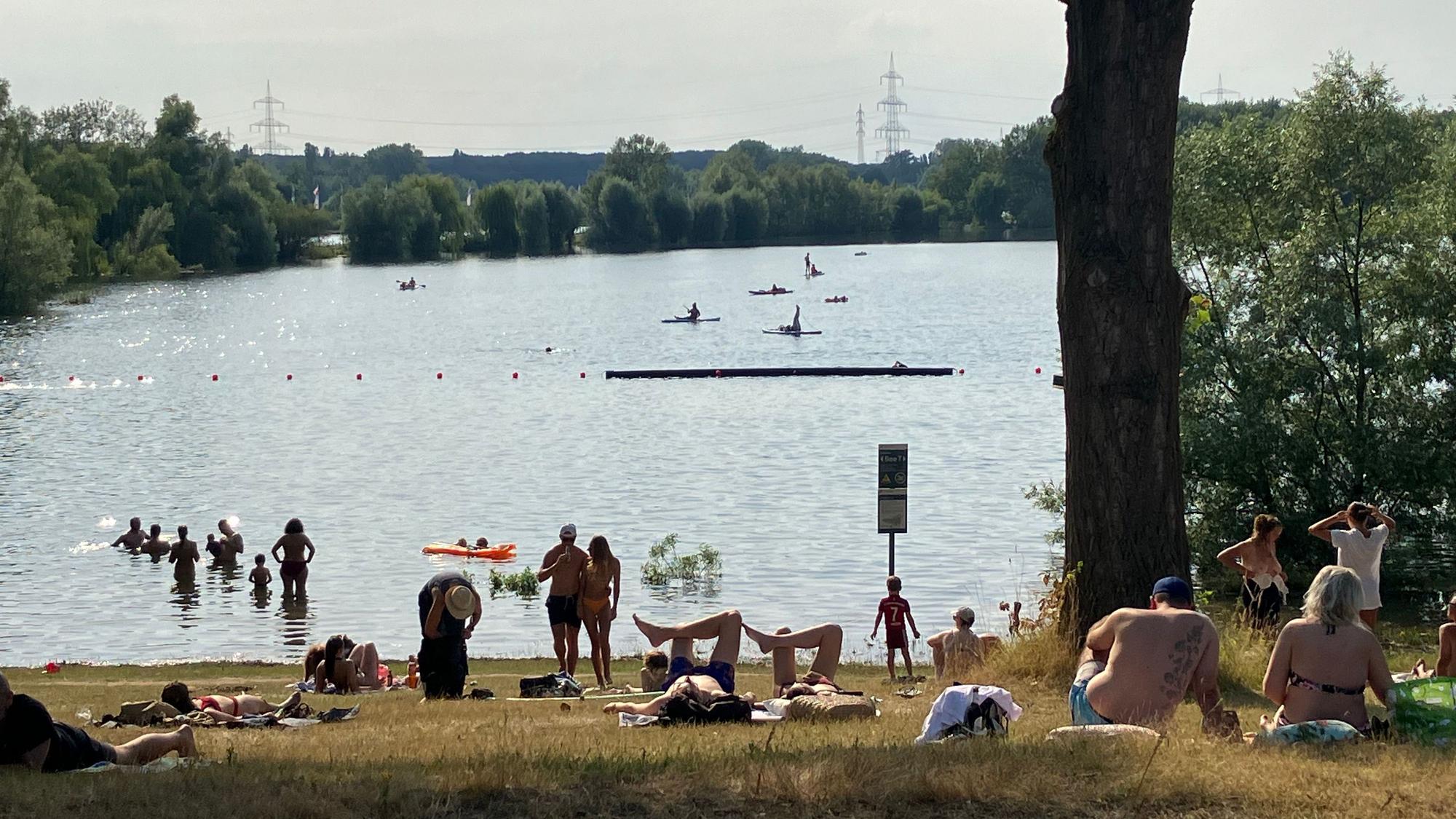 Badegäste sonnen sich an einem Seeufer. Andere schwimmen im Wasser.