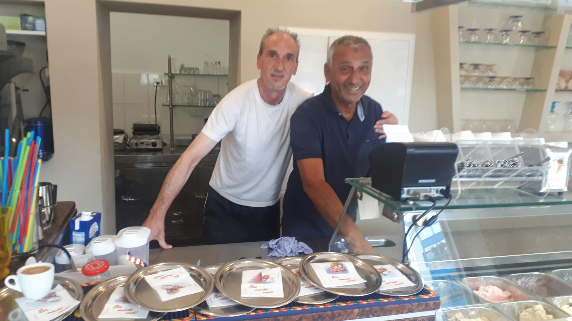 zwei Männer stehen Arm in Arm hinter dem Tresen einer Eisdiele