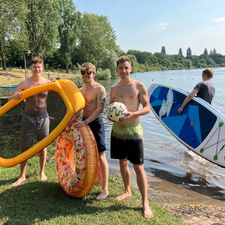 Ruben Neelen (Mitte) und zwei Freunde, alle junge Männer, posieren mit Schimmring und Luftmatratze am Ufer.