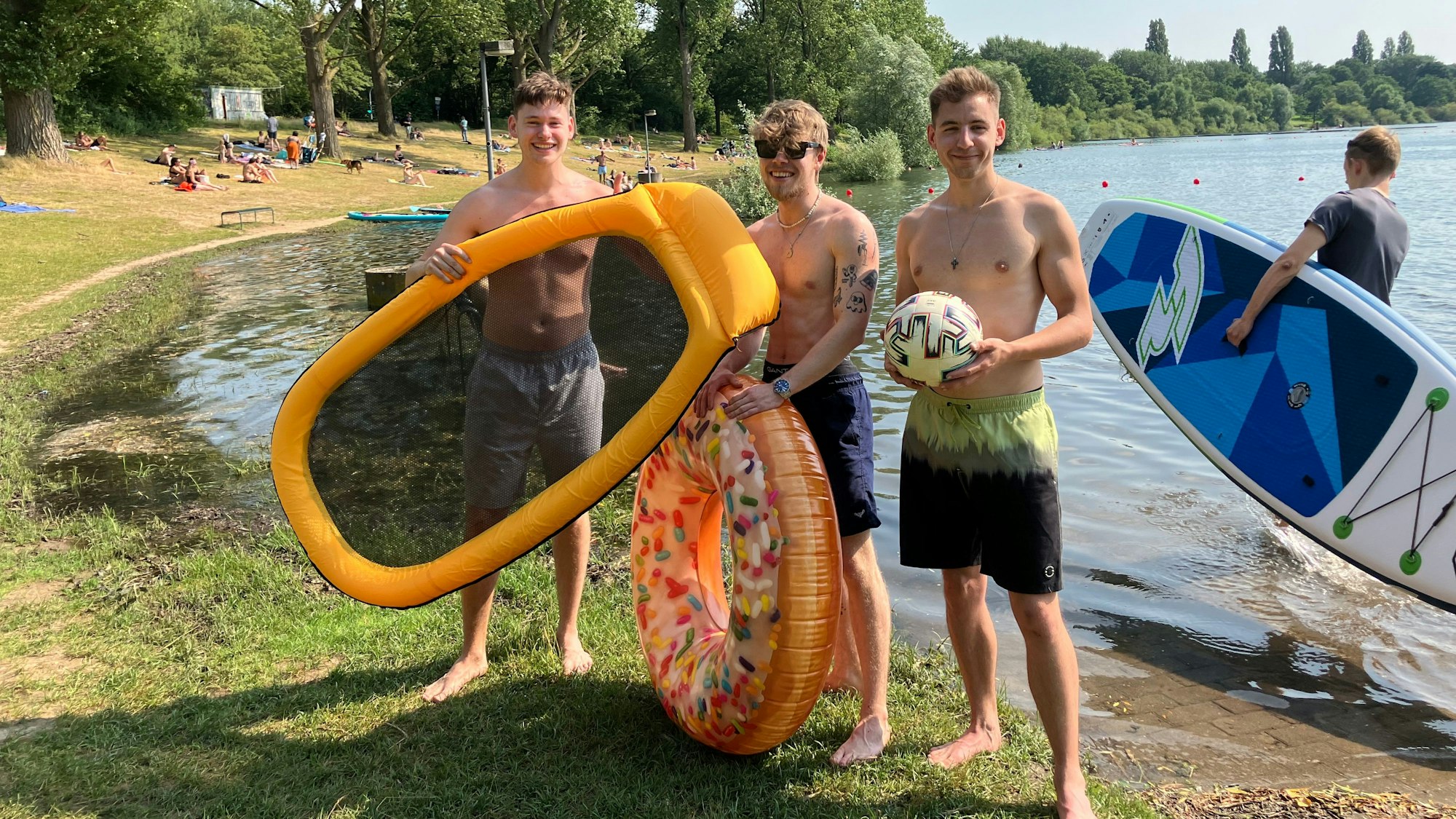 Ruben Neelen (Mitte) und zwei Freunde, alle junge Männer, posieren mit Schimmring und Luftmatratze am Ufer.