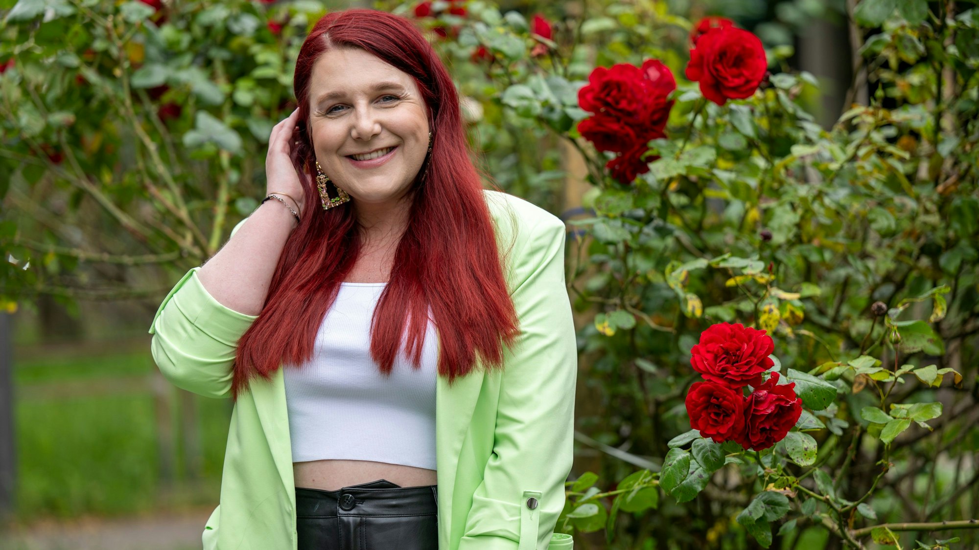 Eine Frau steht lächelnd neben einem Rosenbusch.