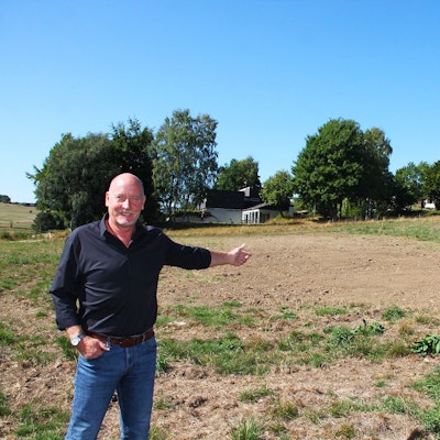Ein Mann in Jeans und schwarzem Hemd zeigt auf eine Wiese, auf der künftig Häuser gebaut werden sollen. Im Hintergrund sind bereits Häuser zu sehen.