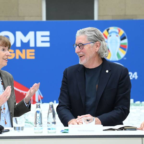 Zu sehen sind V.l. Henriette Reker und Toni Schumacher, die an einem Tisch stehen und lächeln.